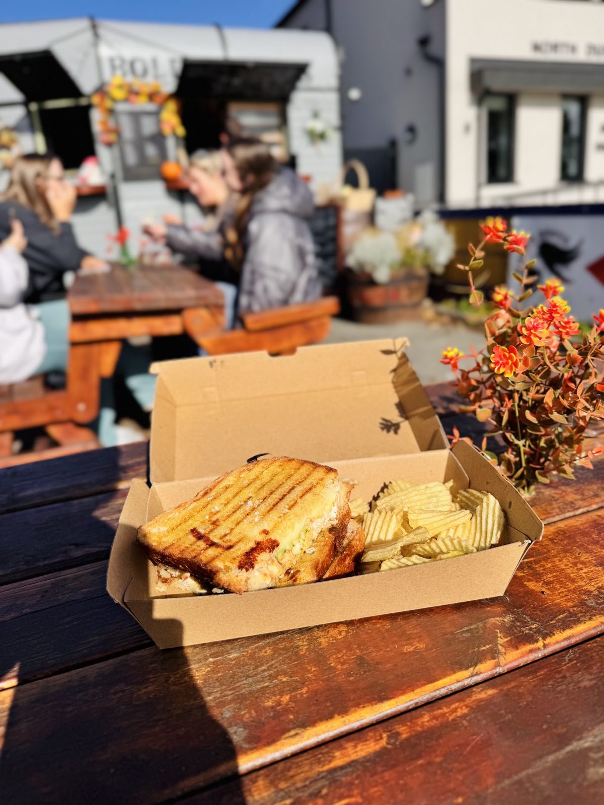 A toasted sandwich and ripple crisps in a kraft box on a timber picnic table, with the trailer behind.