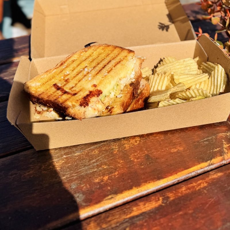 A toasted sandwich with ripple crisps in an open kraft box on a timber table.